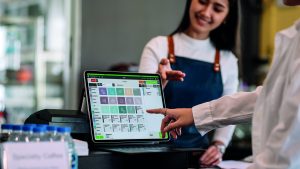 Kellnerin tippt lächelnd auf ein Tablet mit der Gastronovi-Kassenoberfläche, während im Vordergrund eine Hand auf ein Menü-Icon zeigt.